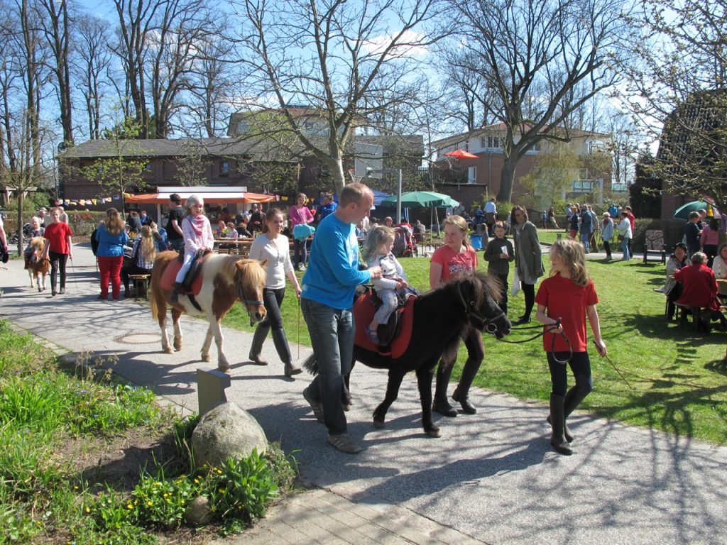 Ein Vater führt sein Kind auf einem kleinen Pony, während mehrere Kinder auf Ponys reiten und von Erwachsenen begleitet werden. Im Hintergrund sind weitere Familien, Spaziergänger und ein kleiner Rummelplatz zu sehen.