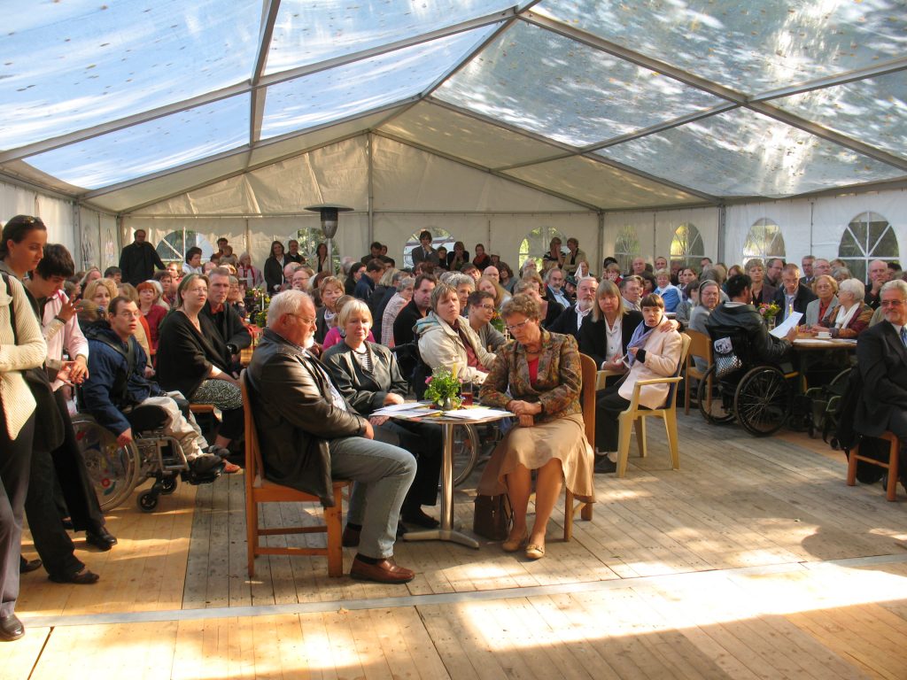 Eine Gruppe von Menschen sitzt unter einem großen Pavillonzelt auf Holzboden. Sie hören aufmerksam zu und richten den Blick nach vorne. Einige Menschen sitzen in Rollstühlen. Im Zelt sind Holztische und -stühle aufgestellt, und es gibt Pflanzen als Dekoration auf den Tischen.