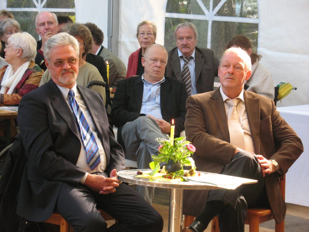 Gruppe von älteren Personen, die in einem Veranstaltungszelt sitzen; zwei Männer im Vordergrund im Anzug, beide mit ernstem Gesichtsausdruck.